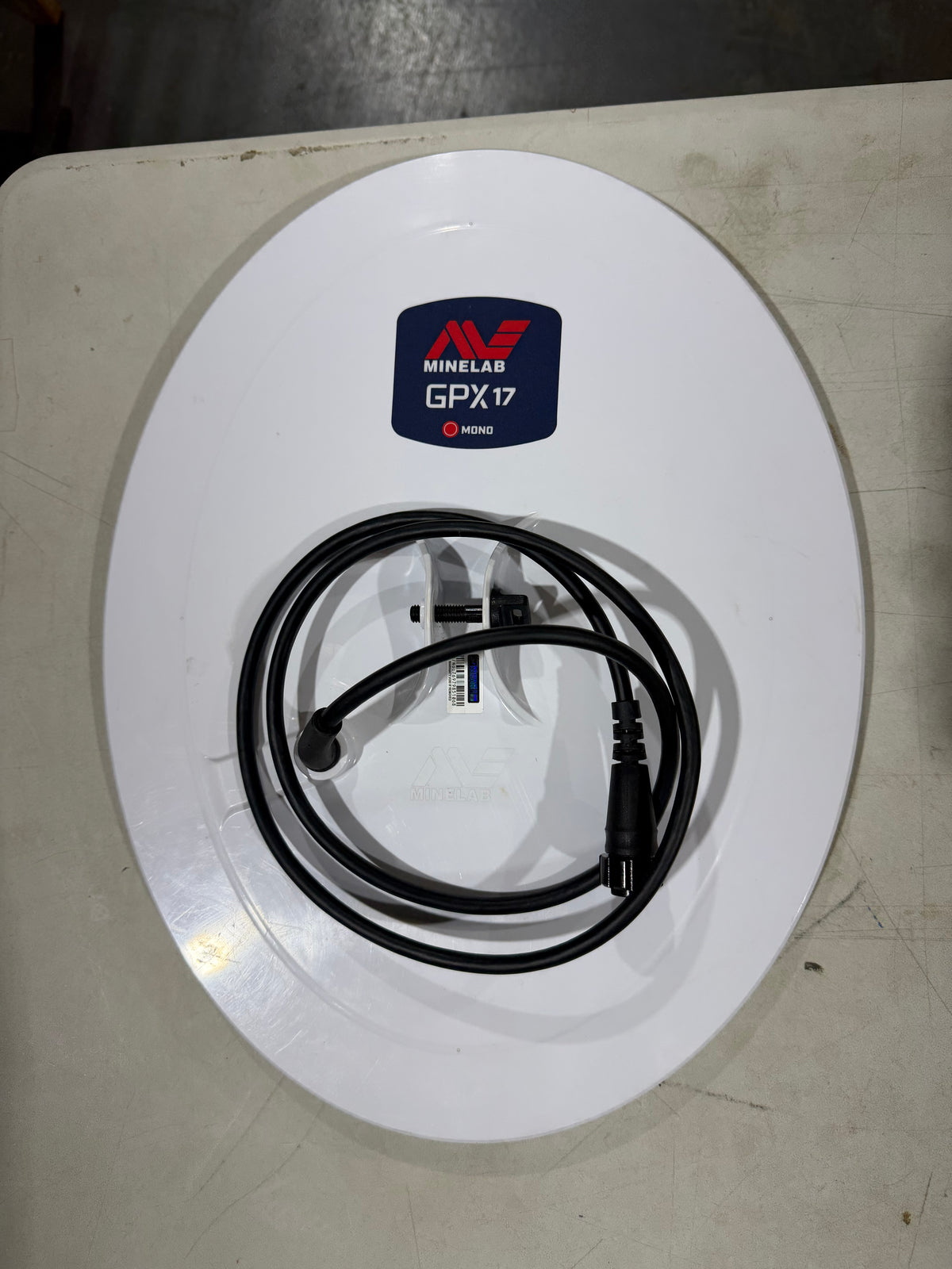 A white oval Minelab GPS antenna labeled 'MINELAB GPX17' with a coiled black power cable, resting on a light gray workbench surface.